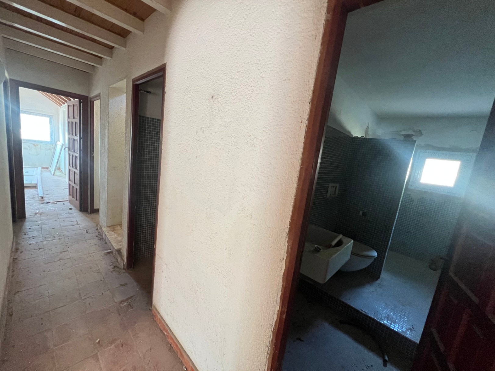 Couloir intérieur avec poutres en bois apparentes, sol en terre cuite et accès à une salle de bain carrelée de mosaïque bleue.