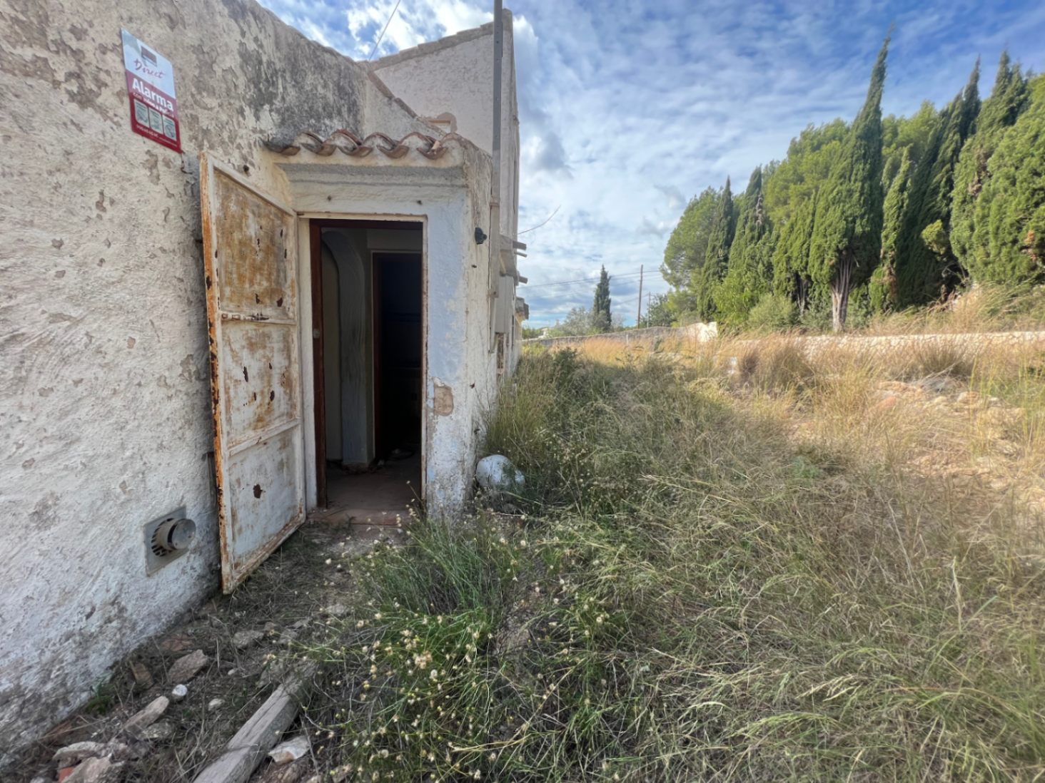 Entrée latérale avec porte métallique rouillée et façade en stuc, entourée d'une végétation sauvage et de cyprès en arrière-plan.