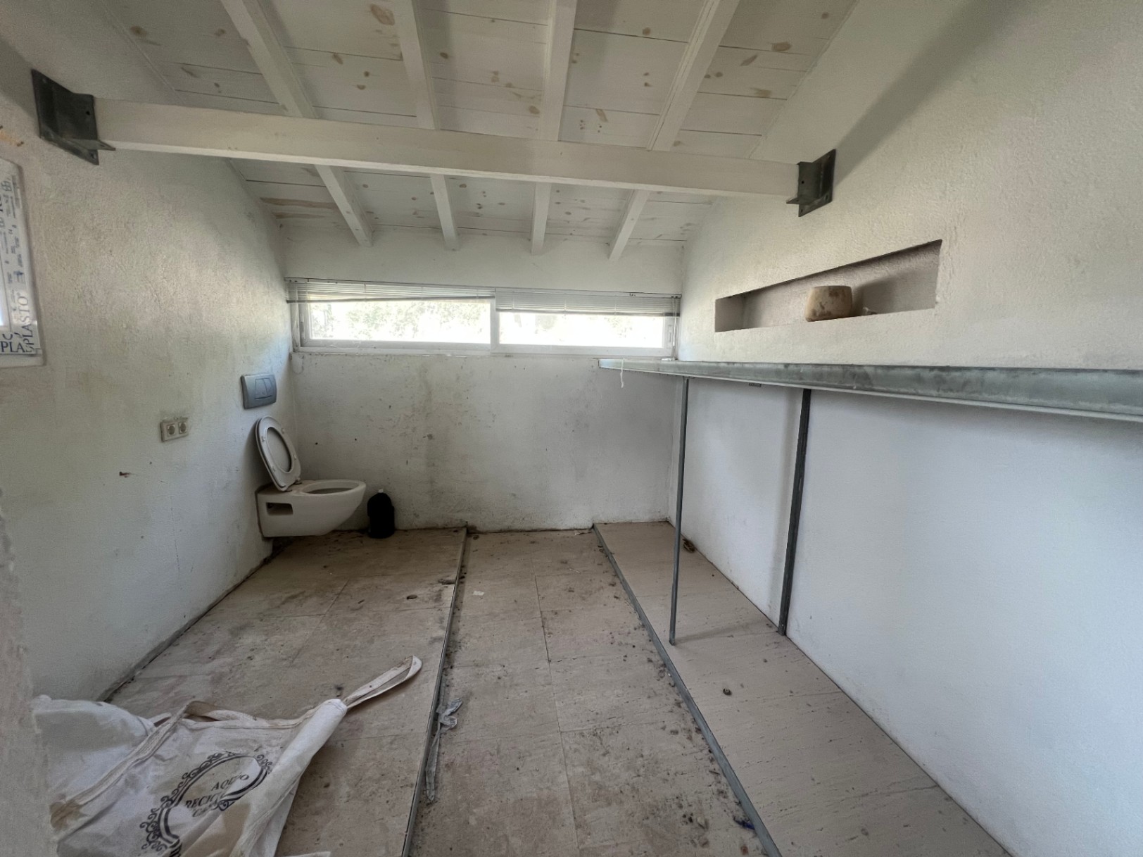 Espace à rénover avec plafond à poutres en bois blanches, fenêtre horizontale, toilettes et étagères métalliques. Idéal pour un futur studio ou débarras.