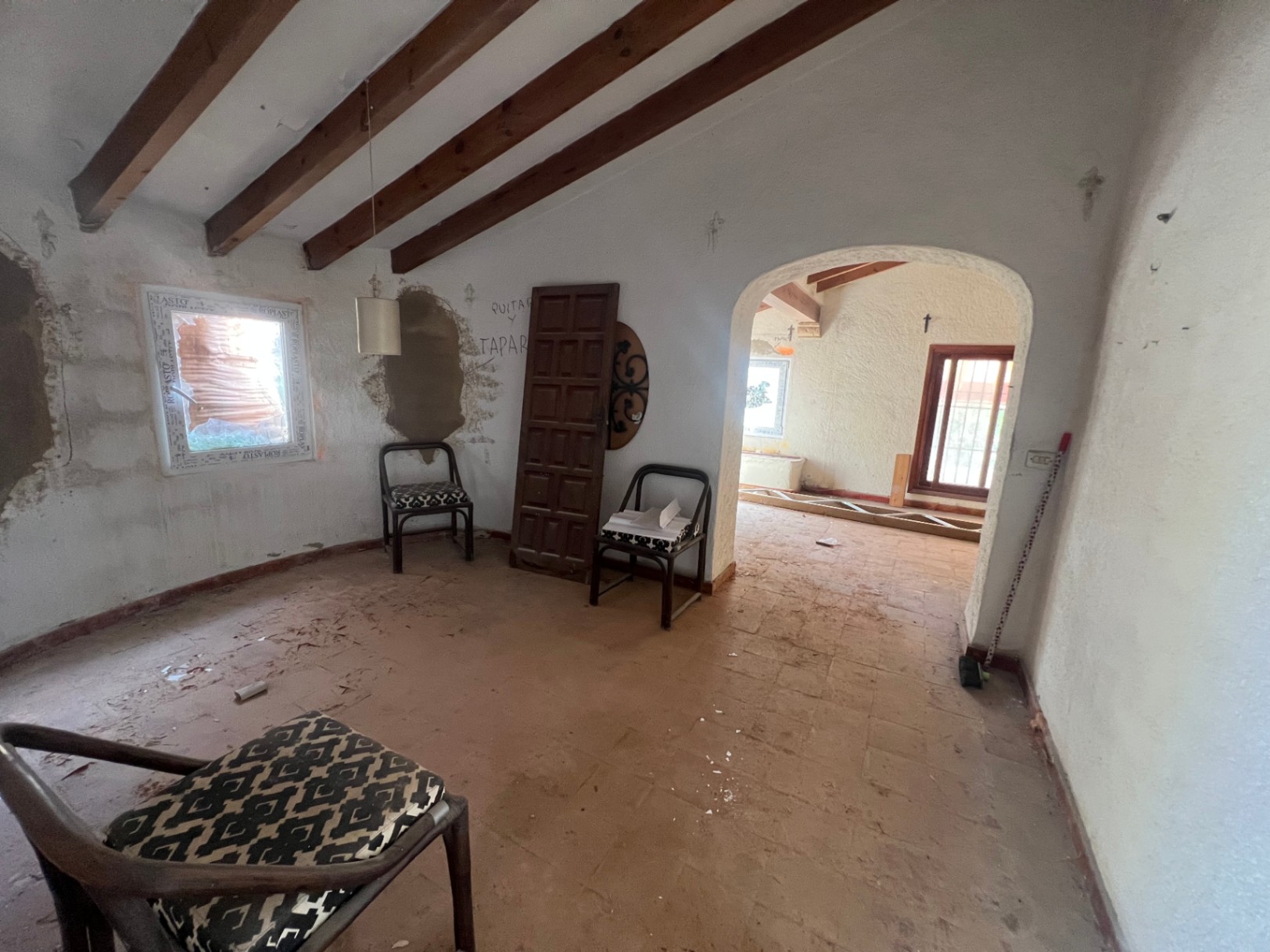 Espace intérieur avec poutres en bois apparentes, sol en terre cuite et une arche décorative. Pièce lumineuse en cours de rénovation offrant un fort potentiel de transformation.