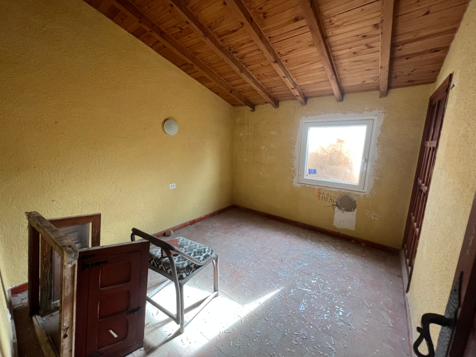 Pièce à rénover avec plafond incliné en poutres de bois apparentes, sol en terre cuite et fenêtre en PVC, offrant un fort potentiel de transformation.