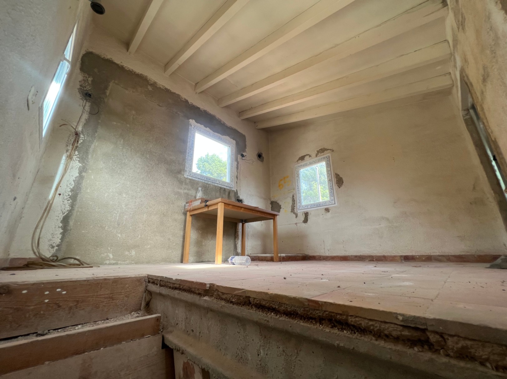 Pièce intérieure en cours de rénovation avec poutres apparentes, sol en terre cuite et murs en béton. Fenêtres à double vitrage et câblage électrique apparent.