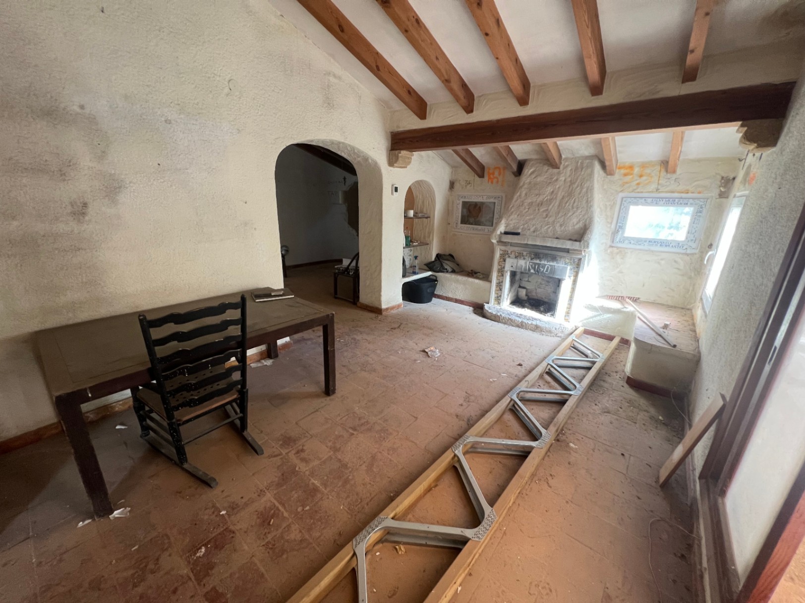 Salon rustique à rénover avec poutres en bois apparentes, cheminée traditionnelle et sol en terre cuite. Des arches ouvrent sur les pièces attenantes.
