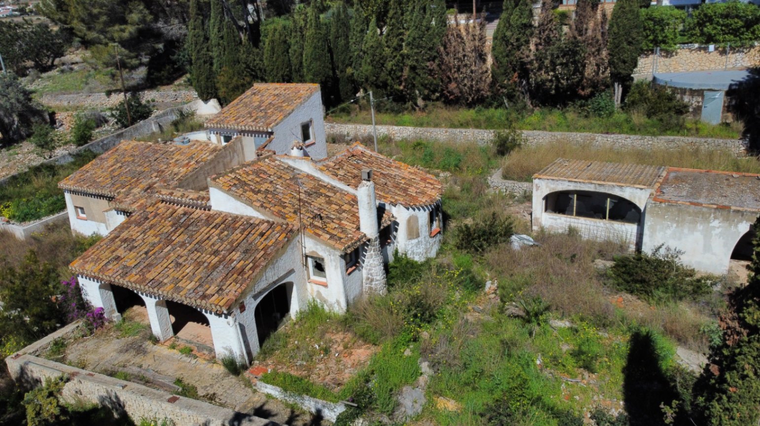 Vue aérienne de la villa avec toitures en tuiles romaines, naya à arches et cheminée en pierre, nichée dans un vaste terrain arboré à rénover.