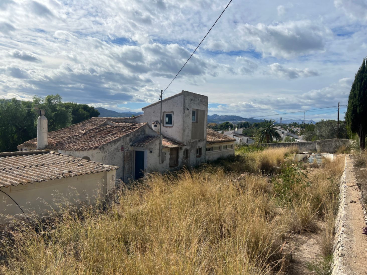 Vue extérieure de la villa avec toitures en tuiles céramiques et façade à rénover, située sur un grand terrain avec végétation sauvage et vue sur les montagnes.