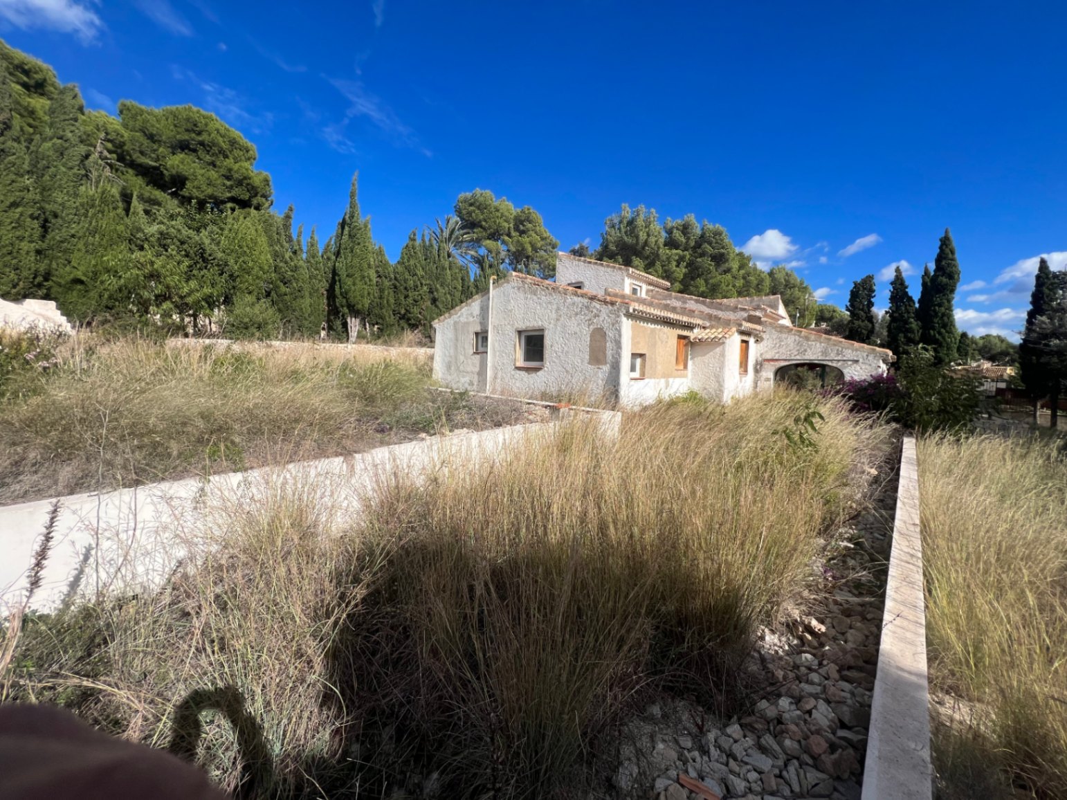 Vue extérieure de la villa traditionnelle avec sa façade texturée et son toit en tuiles, située sur un vaste terrain arboré de pins et de cyprès.