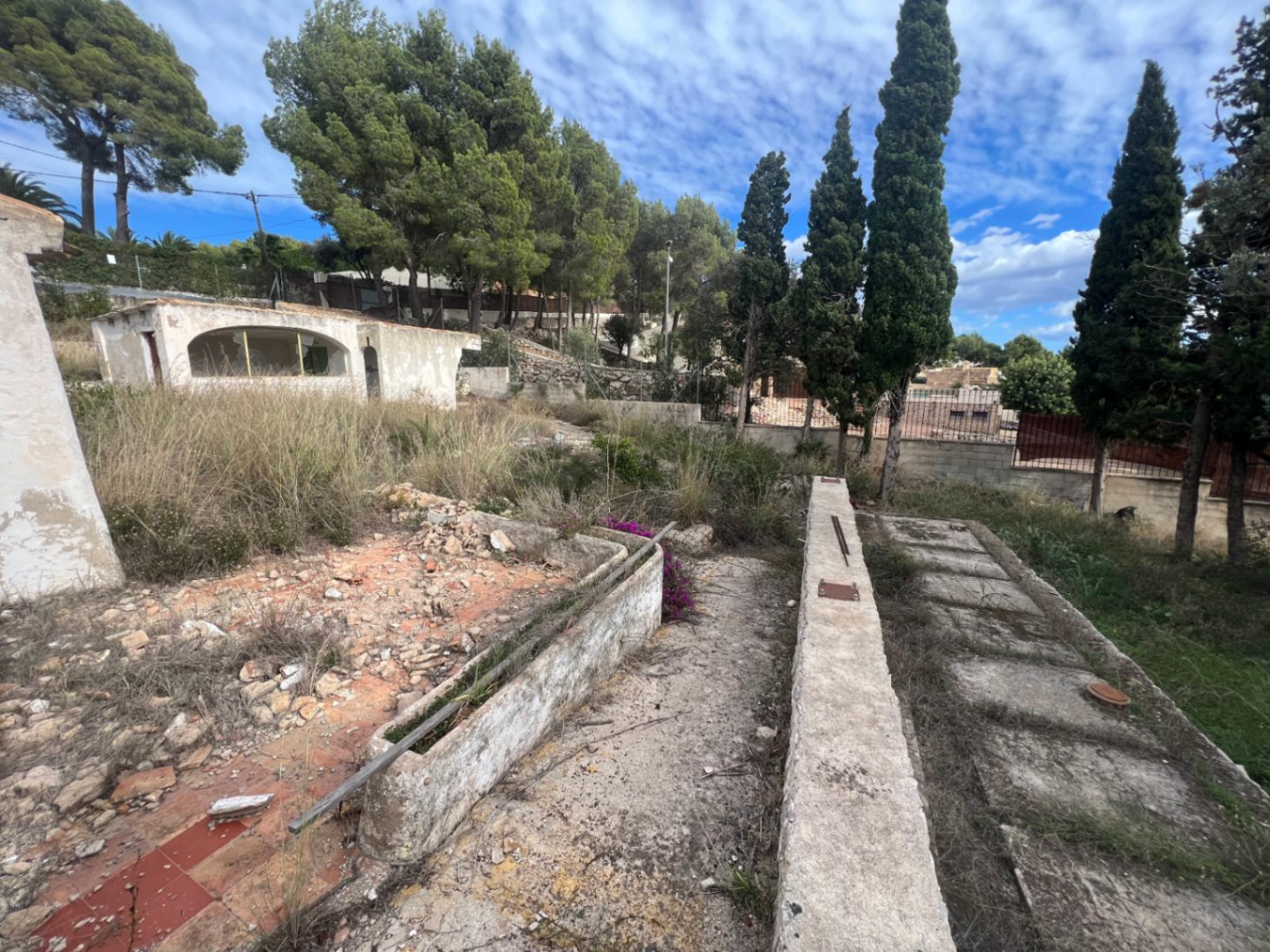 Vue extérieure du terrain avec des structures en béton à rénover, entourée de pins et de cyprès sous un ciel dégagé dans le secteur du Montgó.