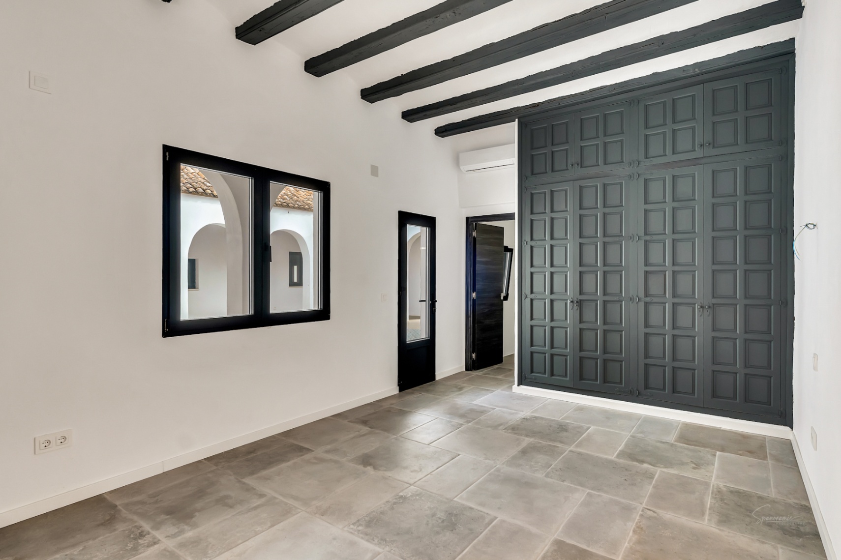 Chambre avec poutres apparentes sombres, sol en pierre naturelle et grand placard intégré. La fenêtre offre une vue sur le patio intérieur aux arcades méditerranéennes.
