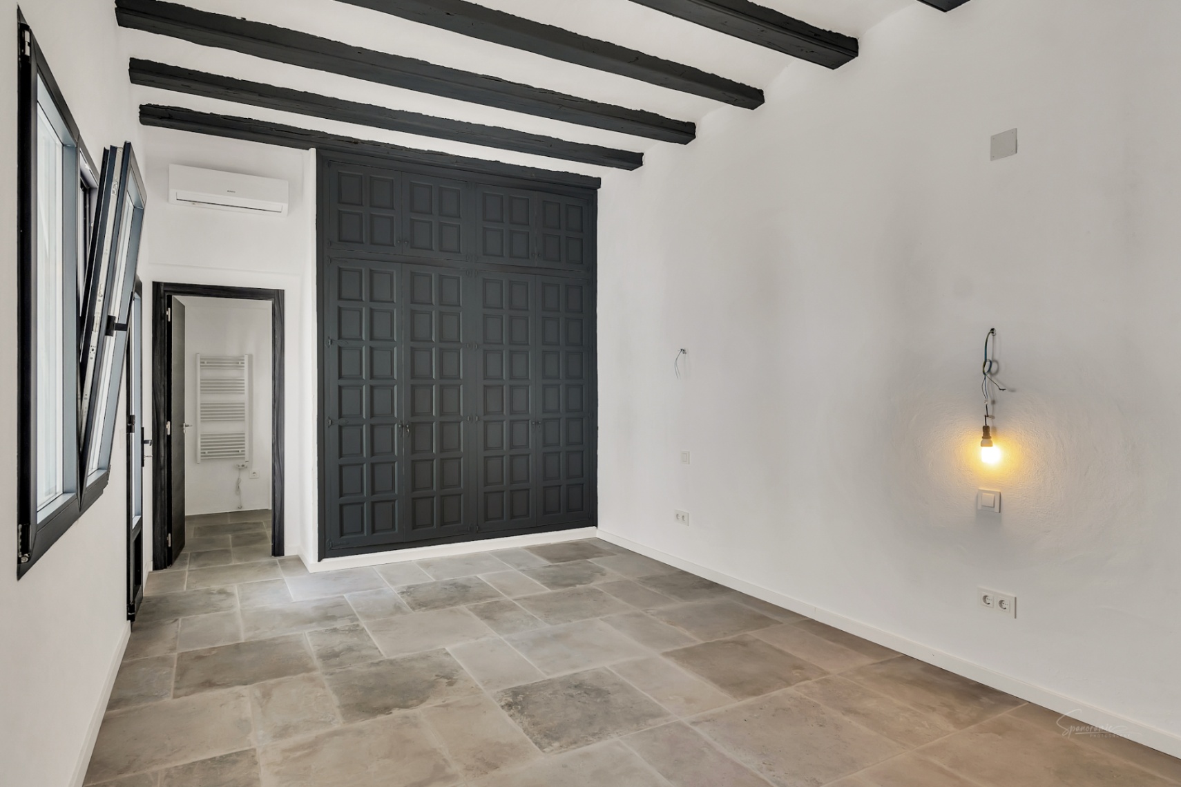 Chambre avec poutres apparentes sombres, sol en pierre naturelle et une grande armoire encastrée noire. Comprend la climatisation et un accès vers la salle d'eau.