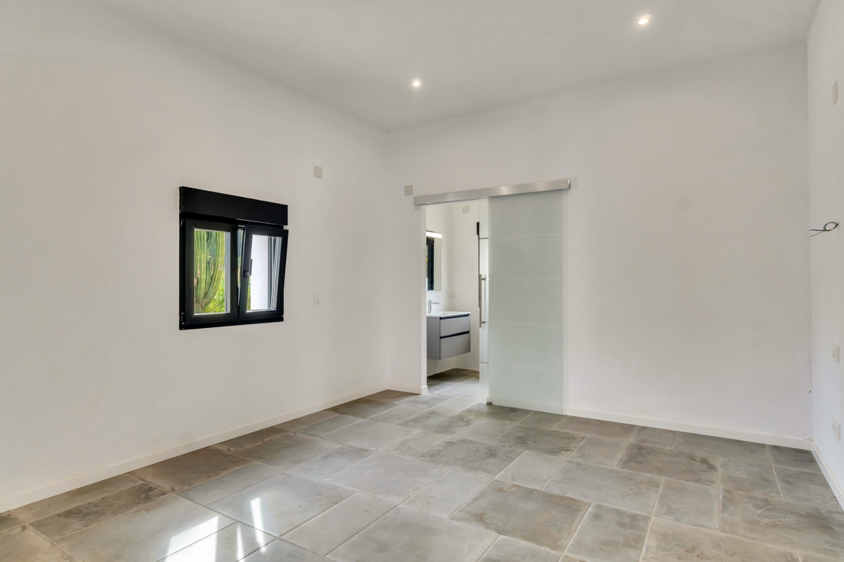 Chambre avec revêtement de sol en pierre, spots encastrés et porte coulissante en verre dépoli ouvrant sur une salle de bain attenante.