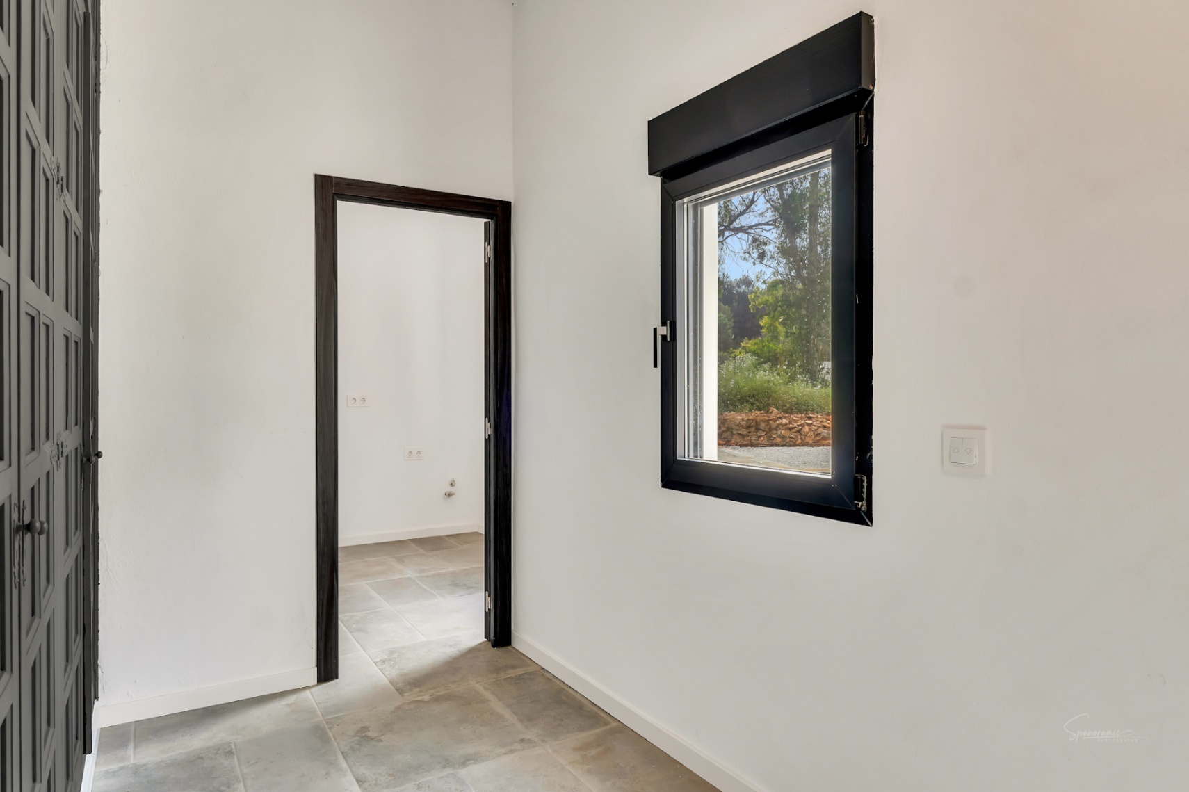 Couloir intérieur avec sol en dalles de pierre et menuiseries sombres. Une fenêtre apporte une lumière naturelle constante et une vue sur la végétation.