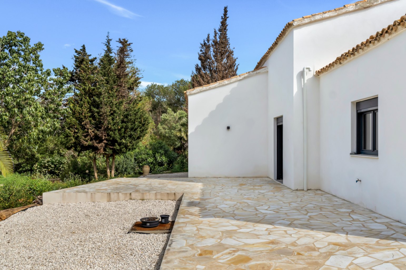 Extérieur de la villa avec façades en stuc blanc et toit en terre cuite, incluant une terrasse en pierre naturelle entourée d'un jardin arboré et de gravier.
