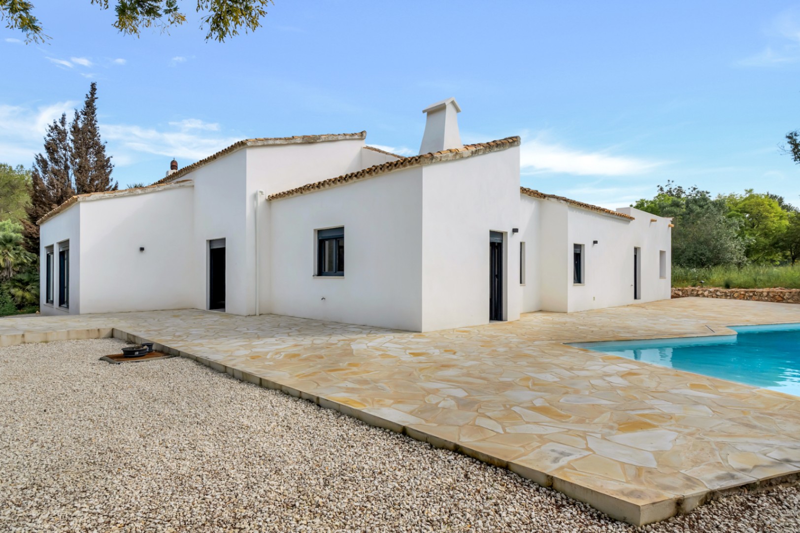 Extérieur de la villa de plain-pied avec murs en stuc blanc, vaste terrasse en pierres irrégulières et coin piscine.