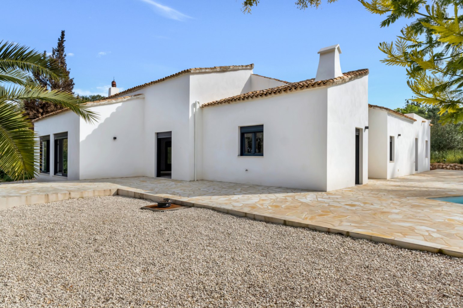 Façade extérieure de style méditerranéen avec murs en stuc blanc, toiture en tuiles et une vaste terrasse en pierre naturelle bordant une zone de gravier.