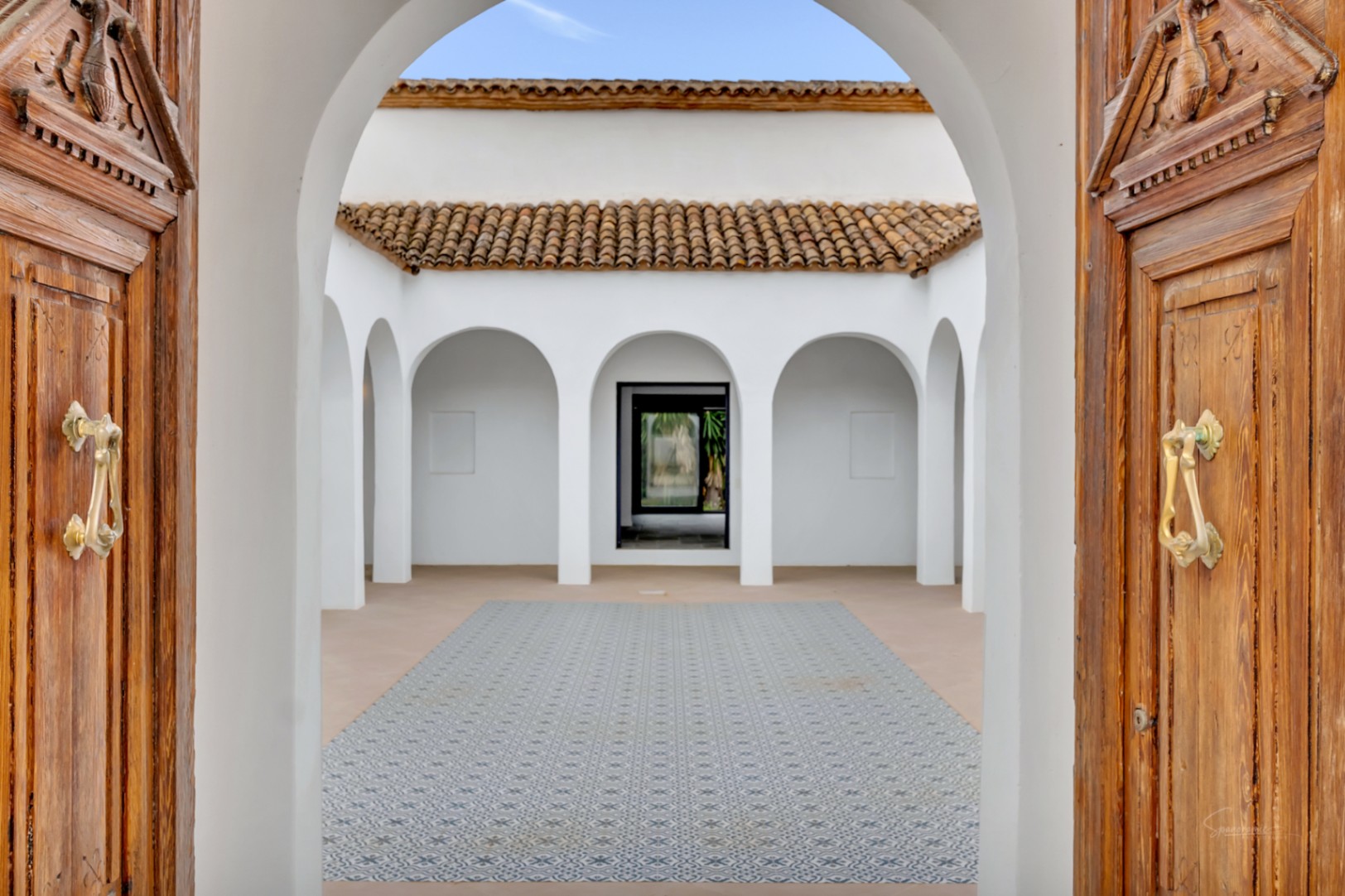 Patio intérieur de style méditerranéen avec arcades blanches, sol en carreaux de ciment décorés et grandes portes en bois sculpté.