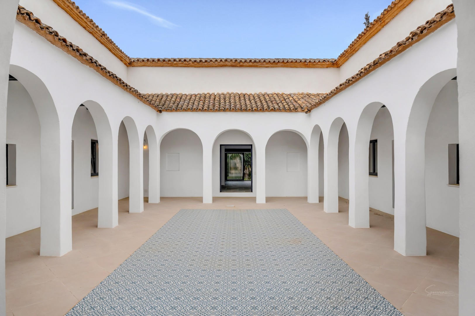 Patio intérieur de style méditerranéen avec des arches blanches symétriques, une toiture en terre cuite et un sol orné de carreaux de ciment aux motifs bleus.