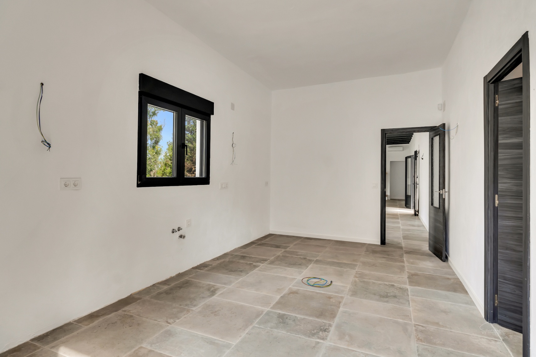 Pièce en cours de rénovation avec sol en grès cérame gris, murs blancs et menuiseries noires. Les portes sombres s'ouvrent sur un couloir dégagé.