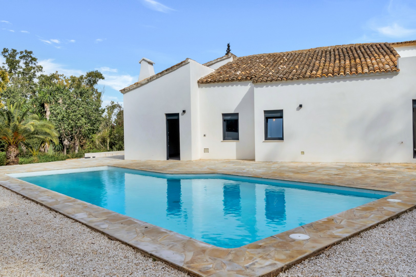 Piscine extérieure entourée d'une terrasse en pierre naturelle et de gravier blanc, bordant une façade méditerranéenne aux tuiles en terre cuite.