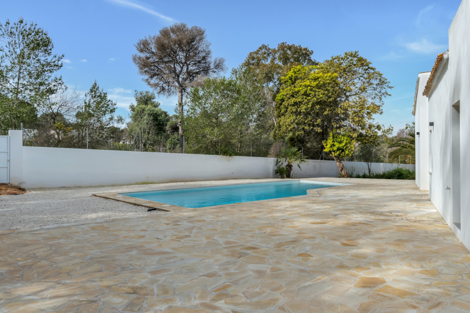 Piscine extérieure entourée d'une vaste terrasse en pierre naturelle et de murs blancs, bordée par une végétation méditerranéenne arborée.