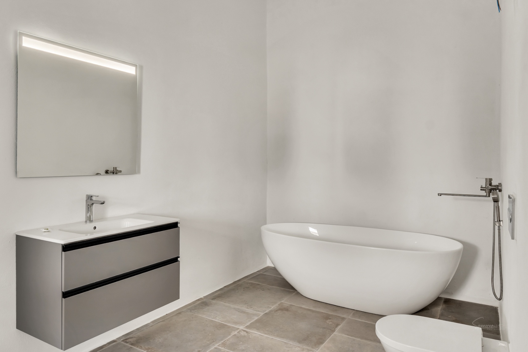 Salle de bain moderne avec baignoire îlot ovale, meuble vasque suspendu gris et sol en pierre naturelle. Un miroir rétroéclairé complète l'esthétique minimaliste.