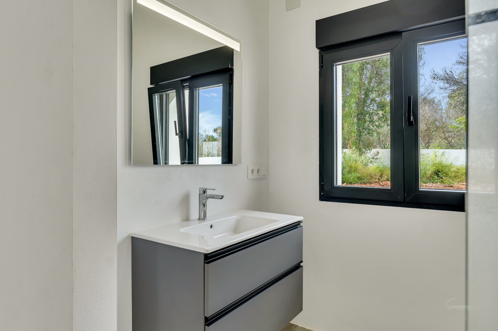 Salle de bain moderne avec meuble vasque gris anthracite, miroir rétroéclairé et fenêtre à cadre noir offrant une vue sur la végétation environnante.