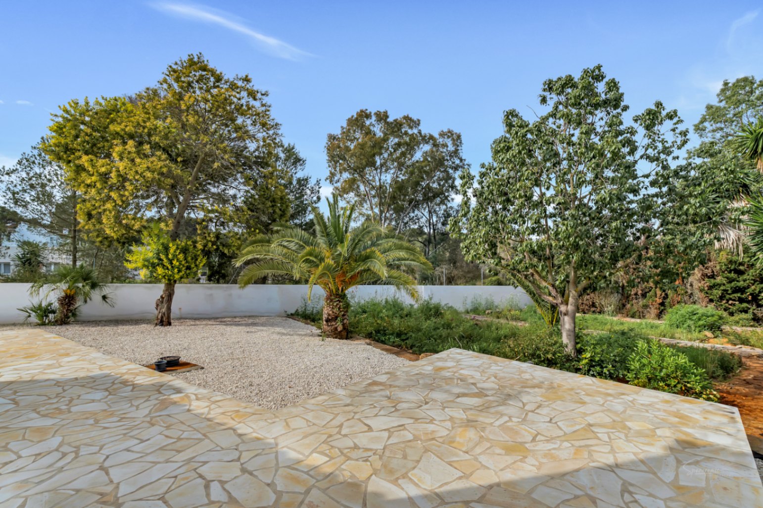 Terrasse extérieure pavée de pierres naturelles irrégulières, s'ouvrant sur un jardin méditerranéen avec palmiers et arbres matures entourés de gravier décoratif.