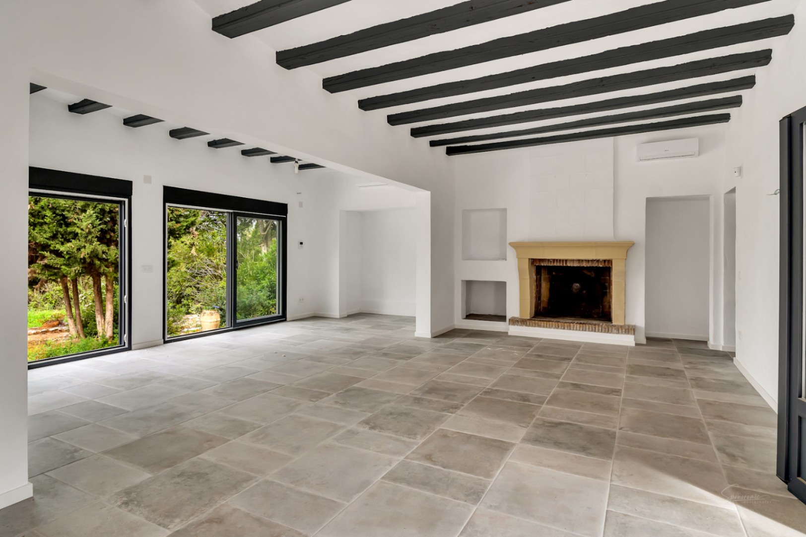 Vaste salon avec cheminée en pierre, poutres apparentes sombres et dalles en pierre naturelle. Les grandes baies vitrées offrent une vue dégagée sur la végétation.
