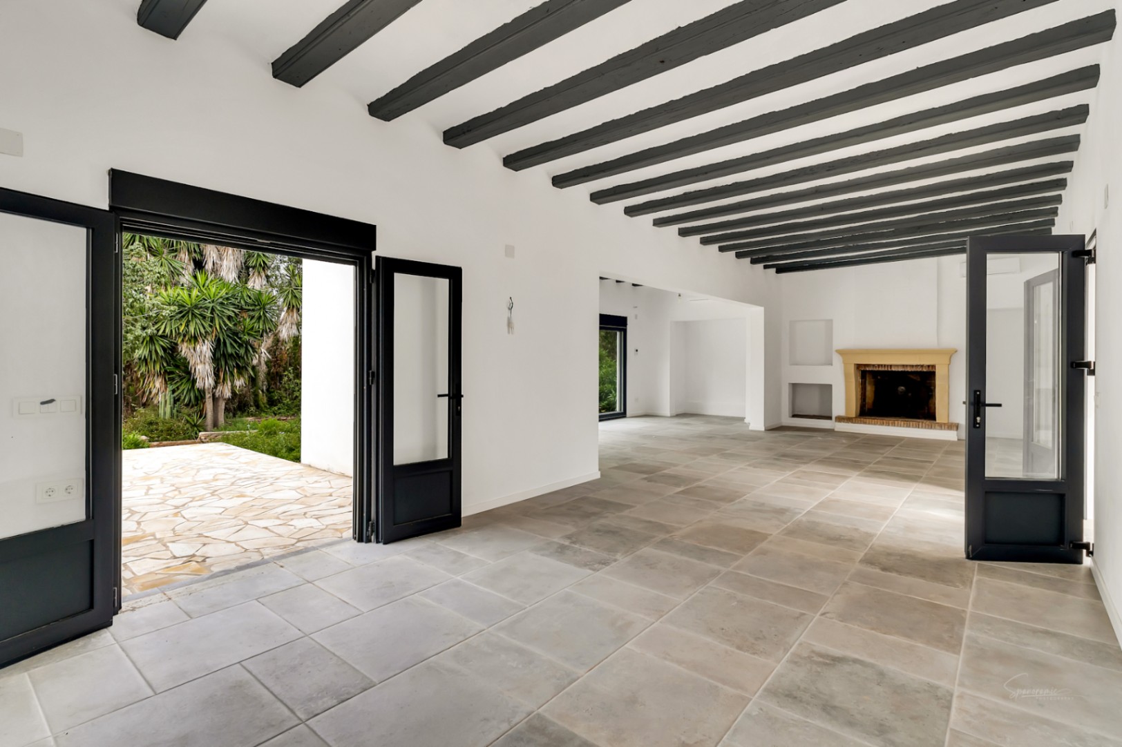 Vaste salon avec poutres apparentes en bois sombre, sol en pierre naturelle et cheminée. De grandes portes vitrées à cadre noir s'ouvrent sur le patio et le jardin méditerranéen.
