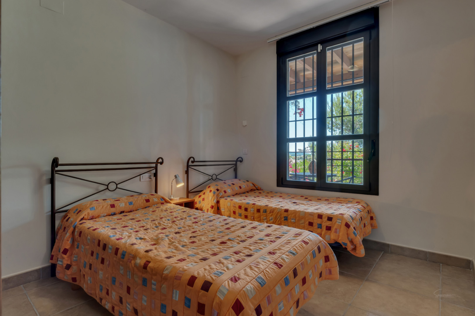 Chambre avec deux lits simples, têtes de lit en fer forgé et sol en grès. La fenêtre encadrée de noir offre une vue sur la végétation extérieure.