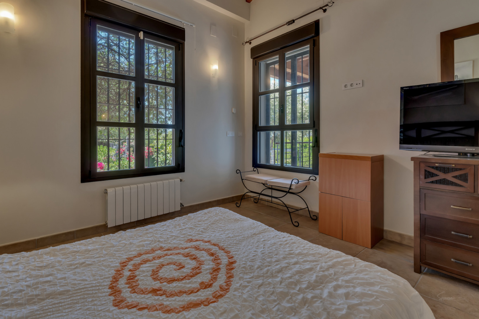 Chambre avec fenêtres à cadre sombre et barreaux, vue sur le jardin, radiateur mural, banc en fer forgé et commode en bois avec télévision.