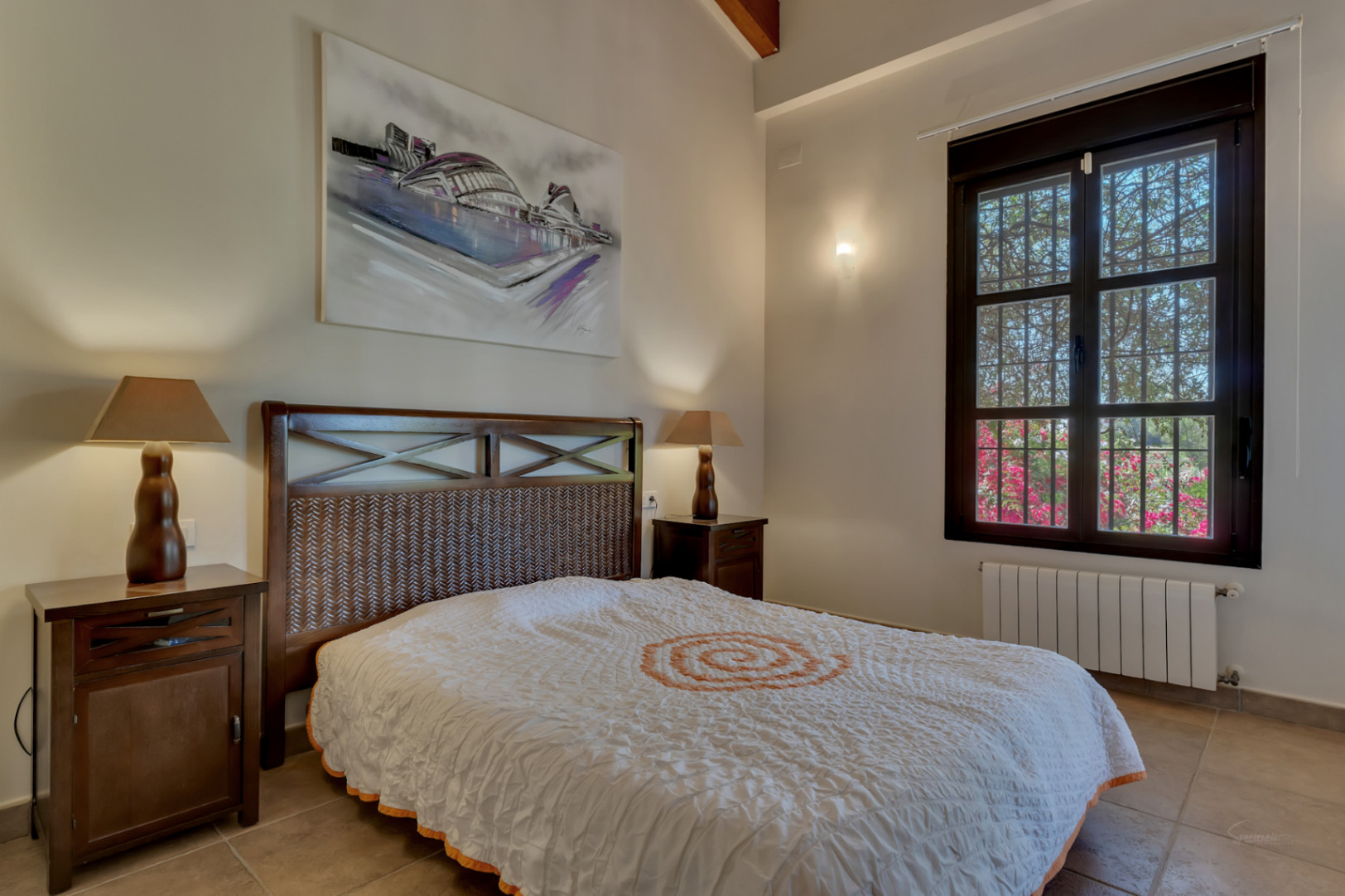 Chambre avec lit en bois sombre et tête de lit tressée, tables de chevet assorties, hauts plafonds avec poutres et fenêtre ouvrant sur le jardin.