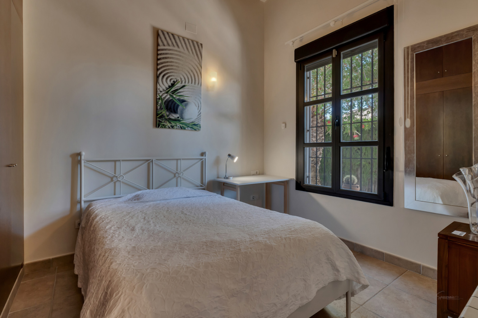 Chambre lumineuse avec lit en métal blanc, coin bureau et fenêtre à cadre sombre offrant une vue sur la végétation du jardin.