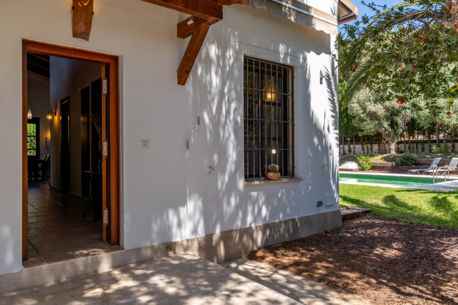 Entrée extérieure avec porte en bois ouverte, façade blanche aux poutres apparentes et vue sur le jardin avec piscine et pelouse ensoleillée.