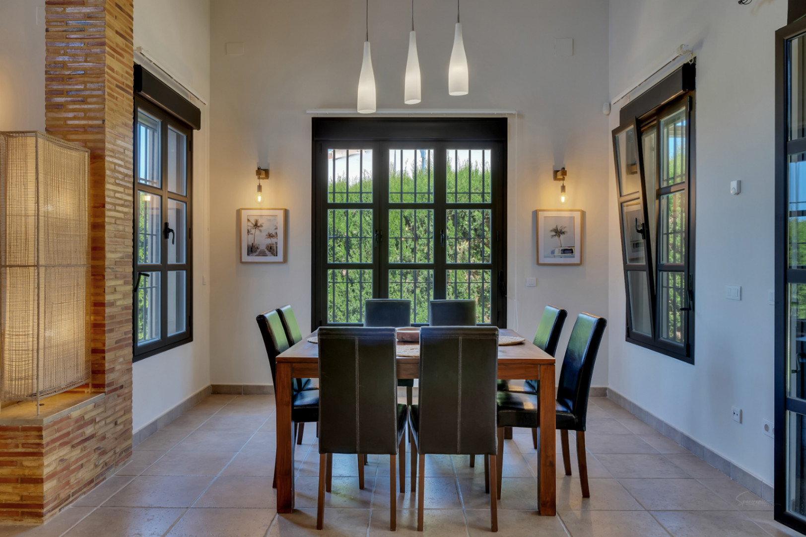 Espace repas avec table en bois et chaises en cuir noir, éclairé par des suspensions triples et des appliques murales près de grandes fenêtres aux cadres noirs.