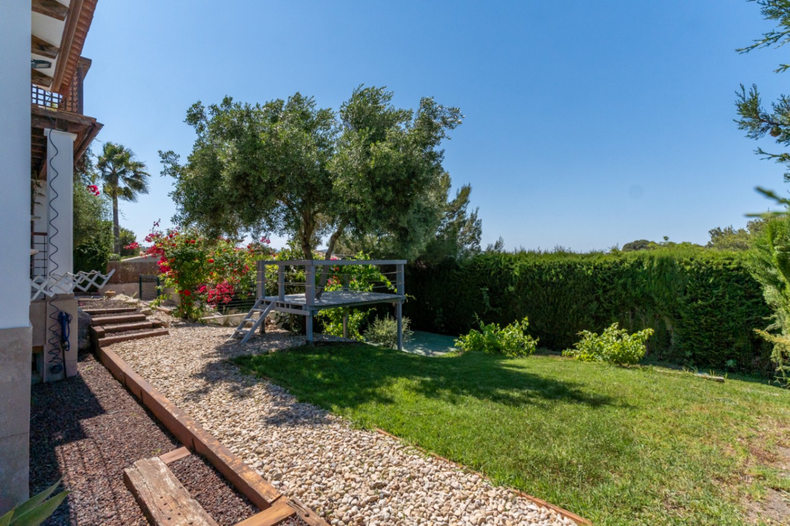 Jardin méditerranéen avec plateforme en bois surélevée sous un olivier, allées en gravier bordées de traverses et hautes haies assurant une intimité totale.