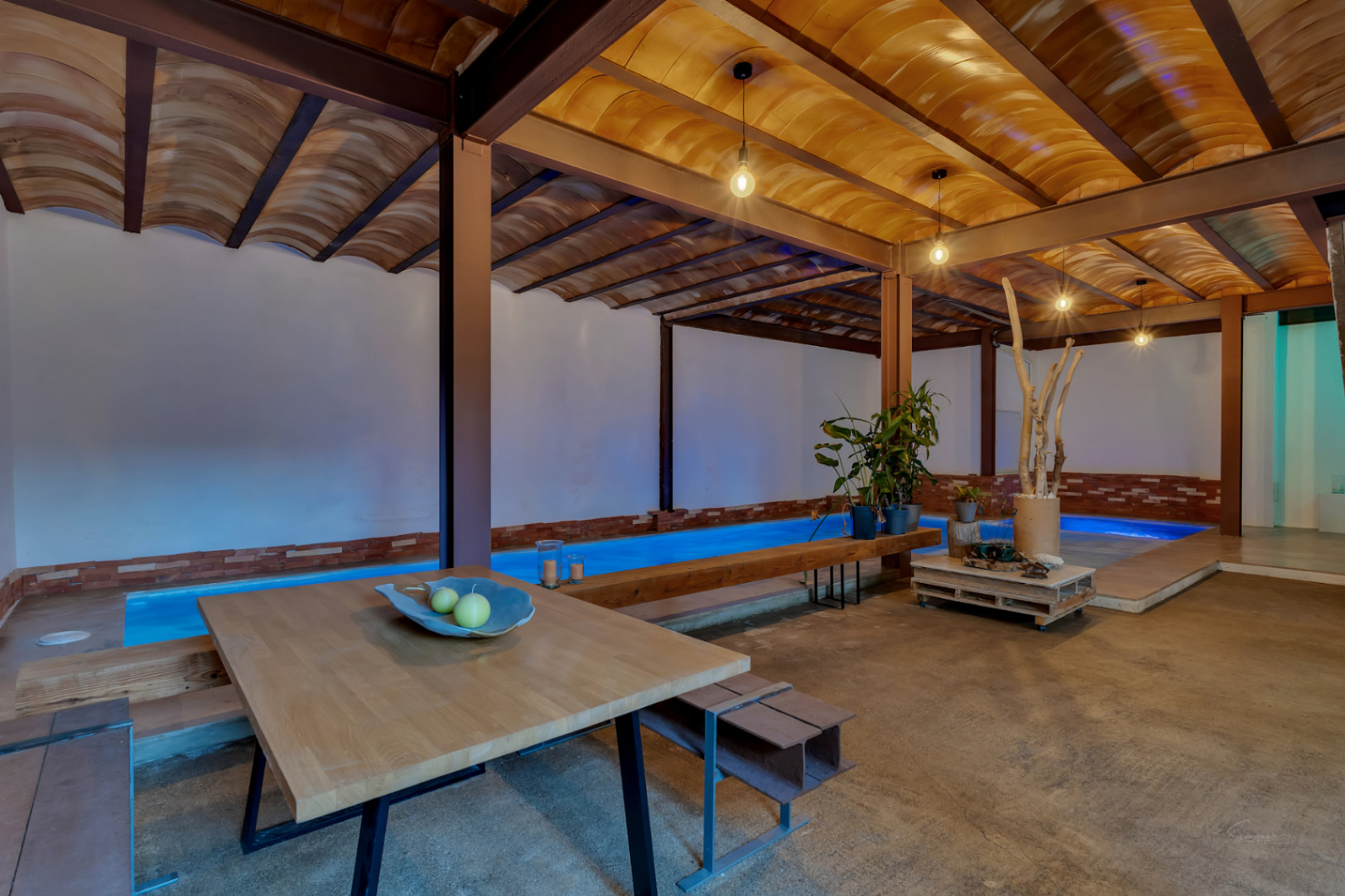 Piscine intérieure chauffée avec éclairage bleu, plafonds voûtés en terre cuite et poutres en acier. Espace ouvert avec coin salon et finitions de style industriel.