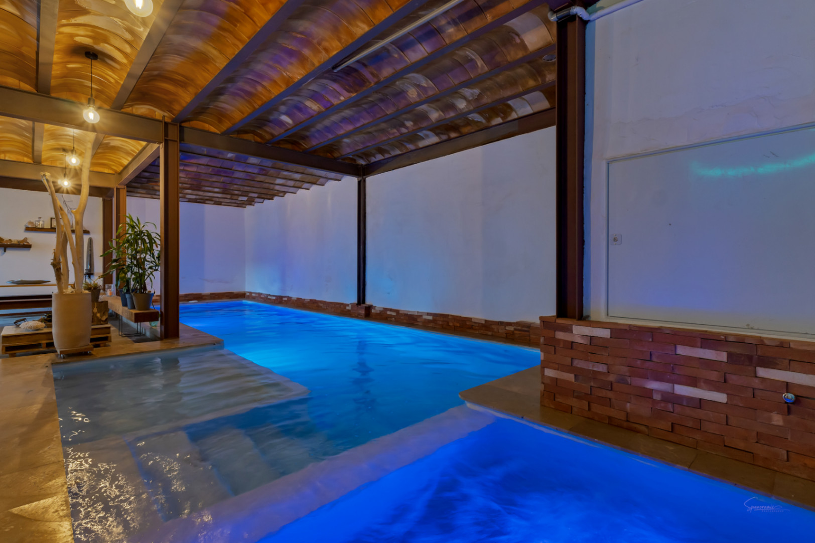 Piscine intérieure chauffée avec éclairage LED bleu, plafonds voûtés en terre cuite et poutres en acier apparentes alliant styles rustique et industriel.