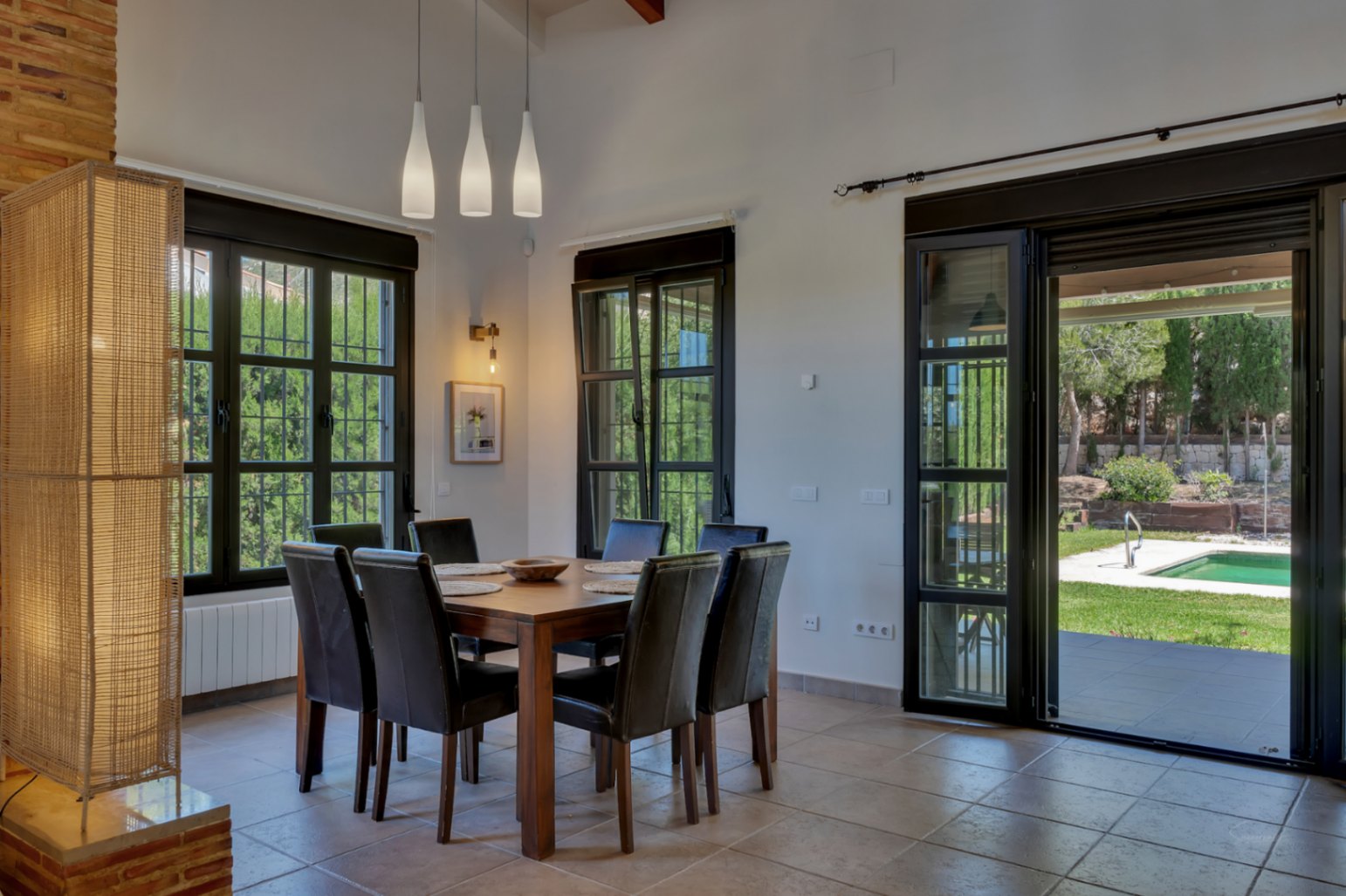 Salle à manger avec table en bois, chaises en cuir et luminaires suspendus, ouverte sur le jardin et la piscine par de larges baies vitrées noires.