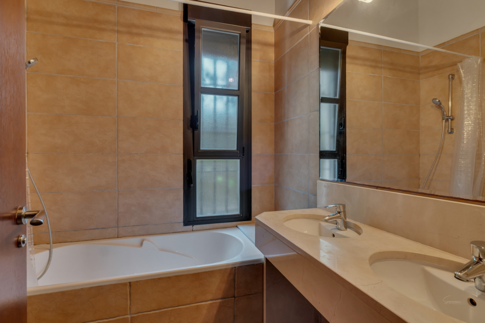 Salle de bain avec double vasque en pierre, baignoire intégrée et carrelage aux tons terreux, éclairée par une fenêtre verticale à cadre sombre.