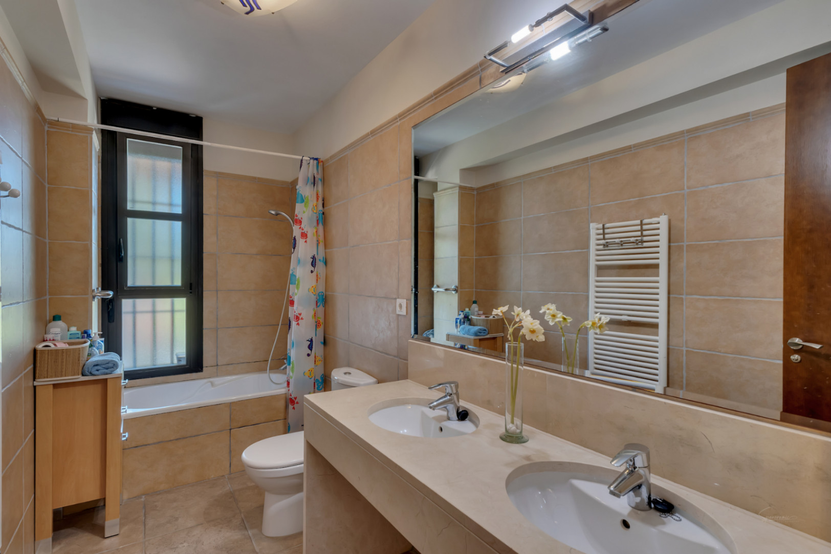 Salle de bain complète avec double vasque, plan de travail en pierre, baignoire intégrée et carrelage mural dans les tons sable chaleureux.