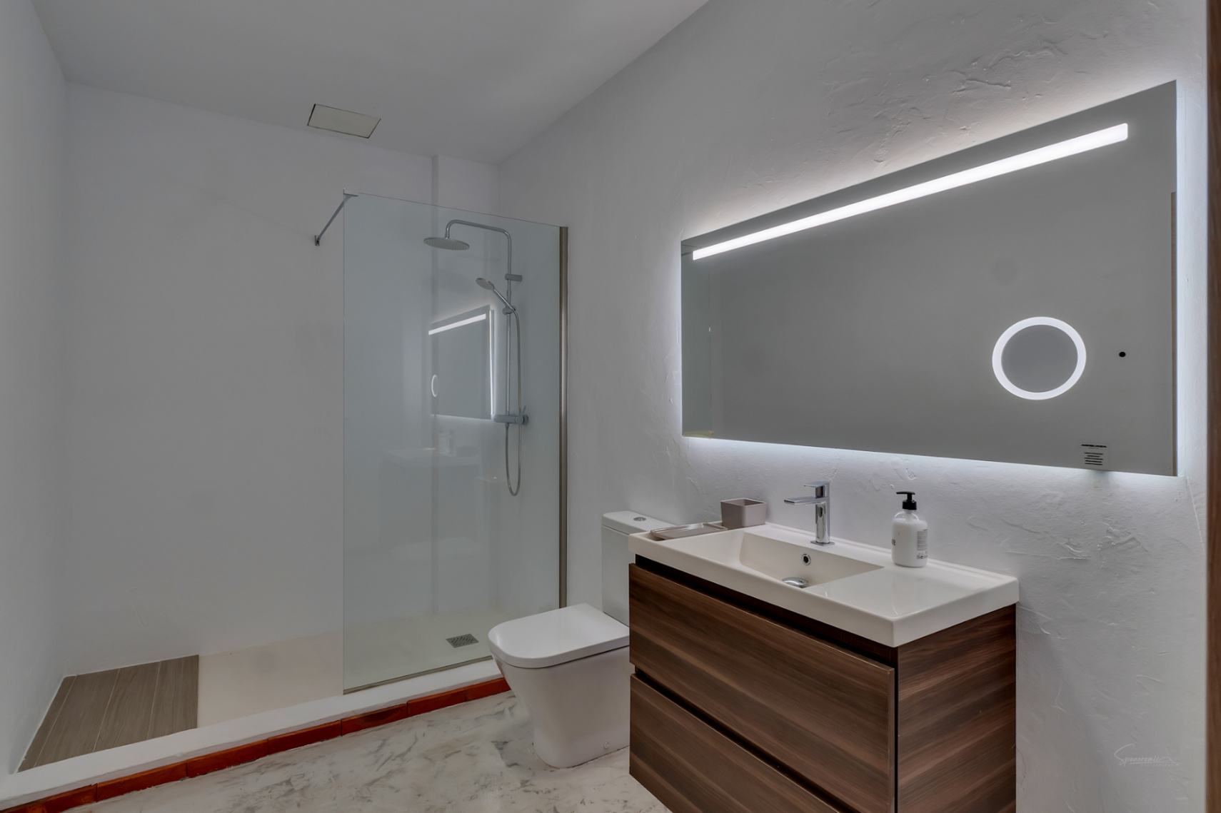 Salle de bains moderne avec meuble vasque suspendu en bois et grand miroir rétroéclairé. Elle dispose d'une douche à l'italienne avec paroi en verre et finitions blanches.