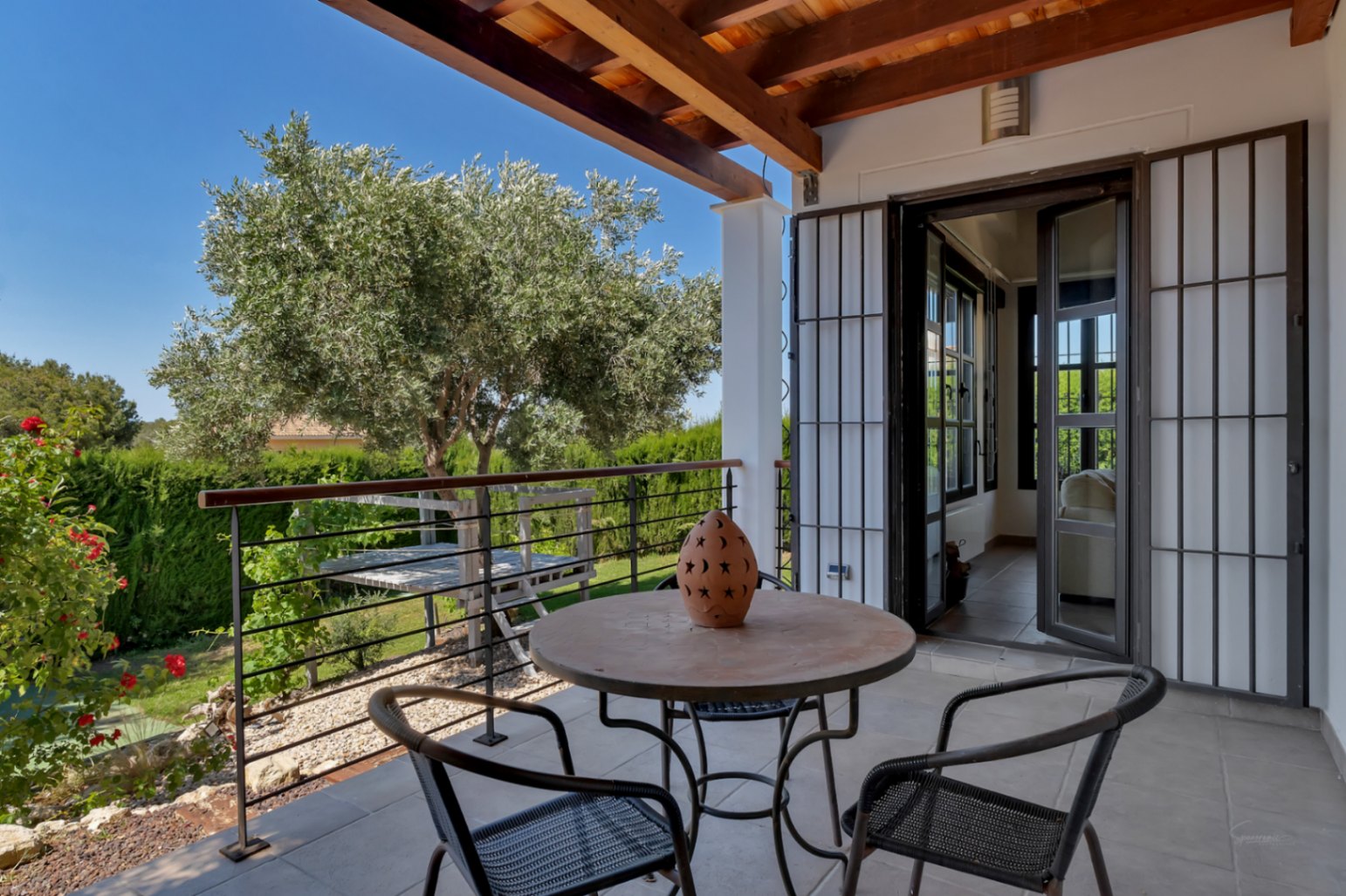 Terrasse couverte avec poutres en bois apparentes, coin salon extérieur et vue sur le jardin paysager agrémenté d'un olivier.