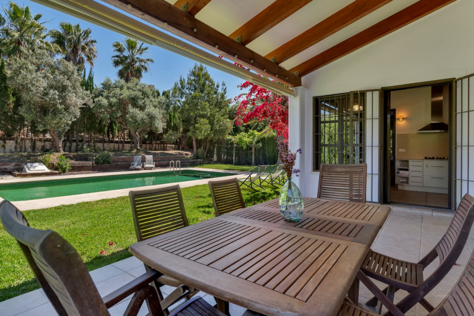 Terrasse couverte avec poutres en bois et coin repas extérieur, offrant une vue sur le jardin méditerranéen et la piscine rectangulaire.