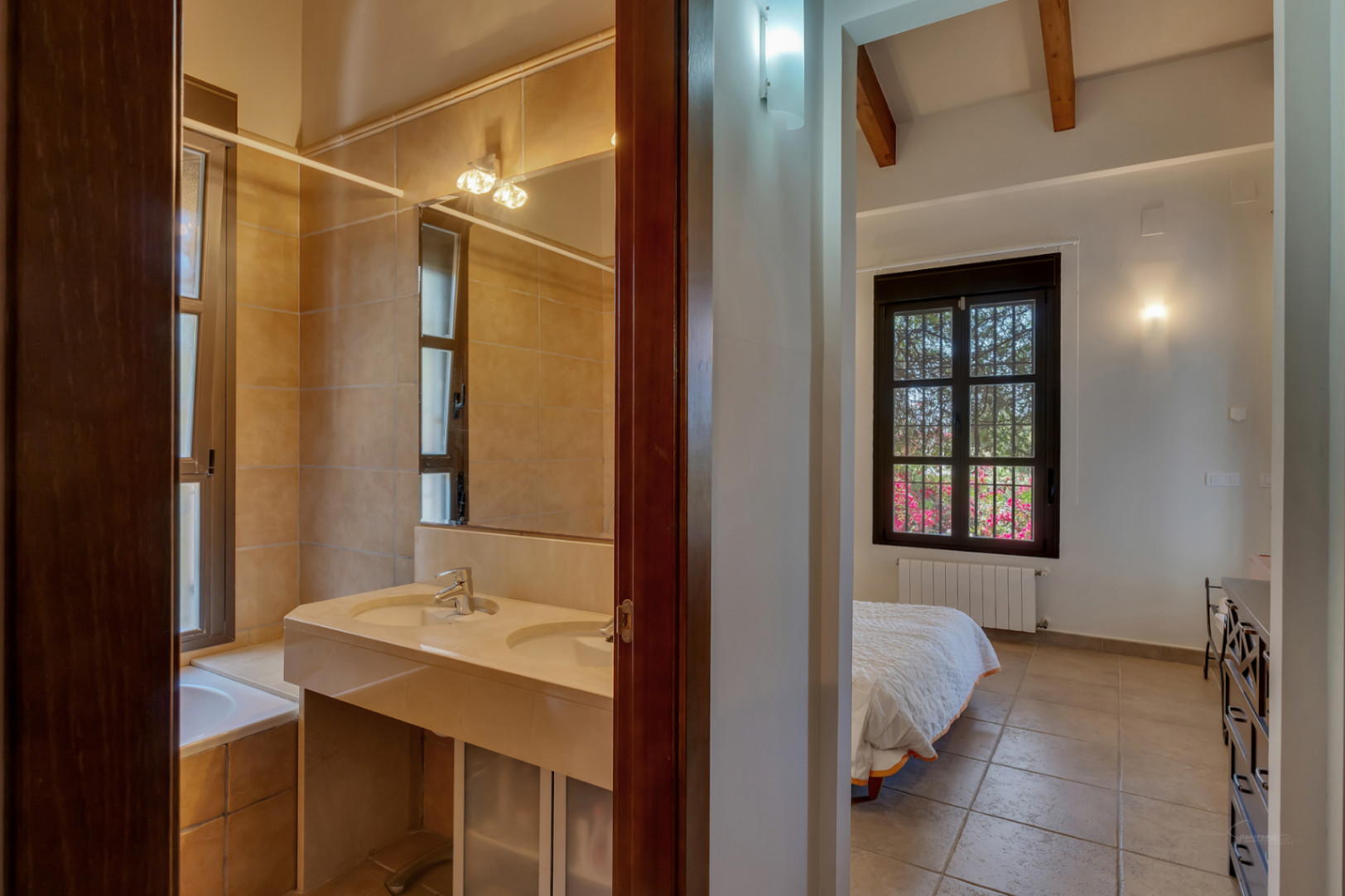 Vue depuis le couloir montrant une salle de bain avec double vasque en pierre et une chambre avec poutres apparentes et vue sur le jardin.