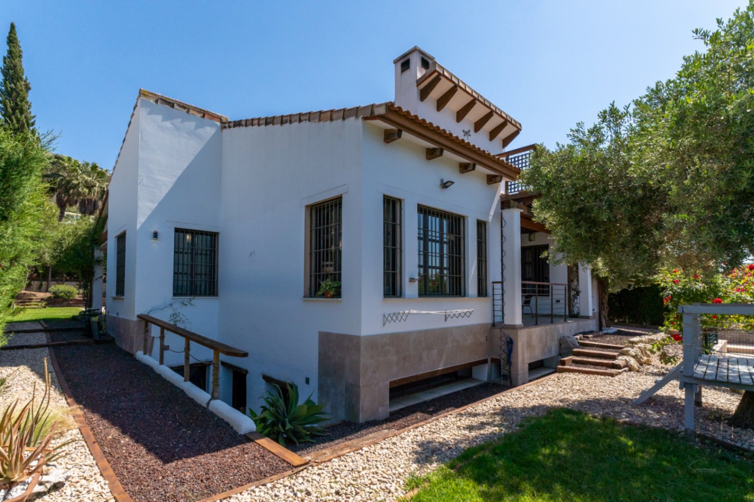 Vue extérieure de la villa avec façade blanche, poutres en bois et soubassement en pierre. Le jardin propose des allées en gravier et un accès au niveau inférieur.