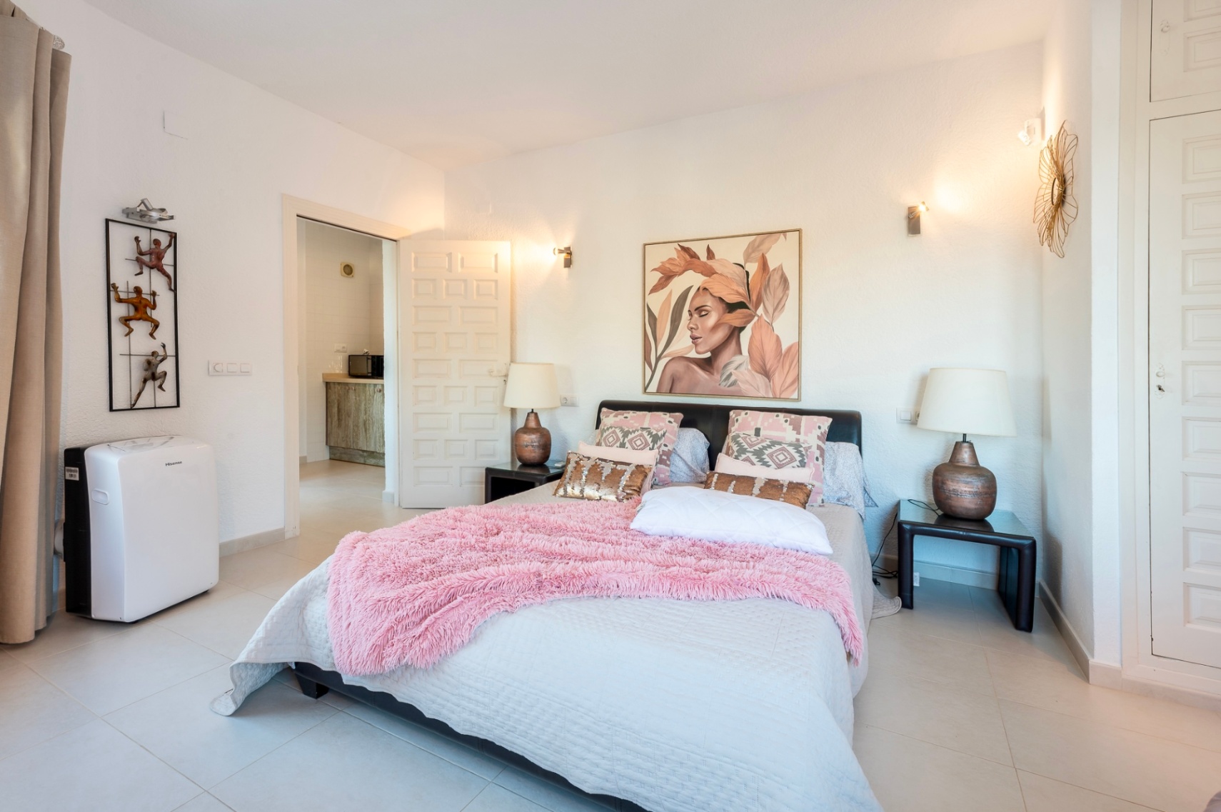 Chambre avec lit double, textiles roses et appliques murales. Elle comprend des placards intégrés blancs, une climatisation portable et un sol en carrelage clair.