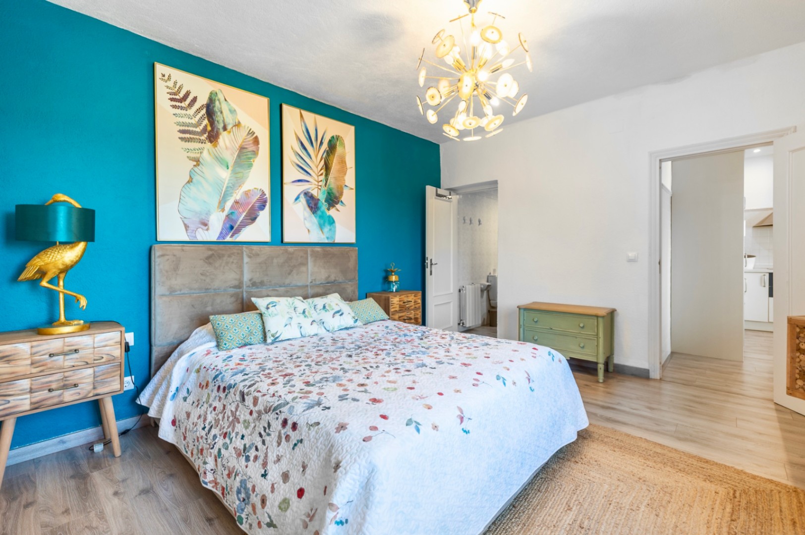 Chambre avec mur d'accent turquoise, tête de lit rembourrée et lustre doré. Comprend une salle de bain attenante, des œuvres botaniques et un sol stratifié clair.