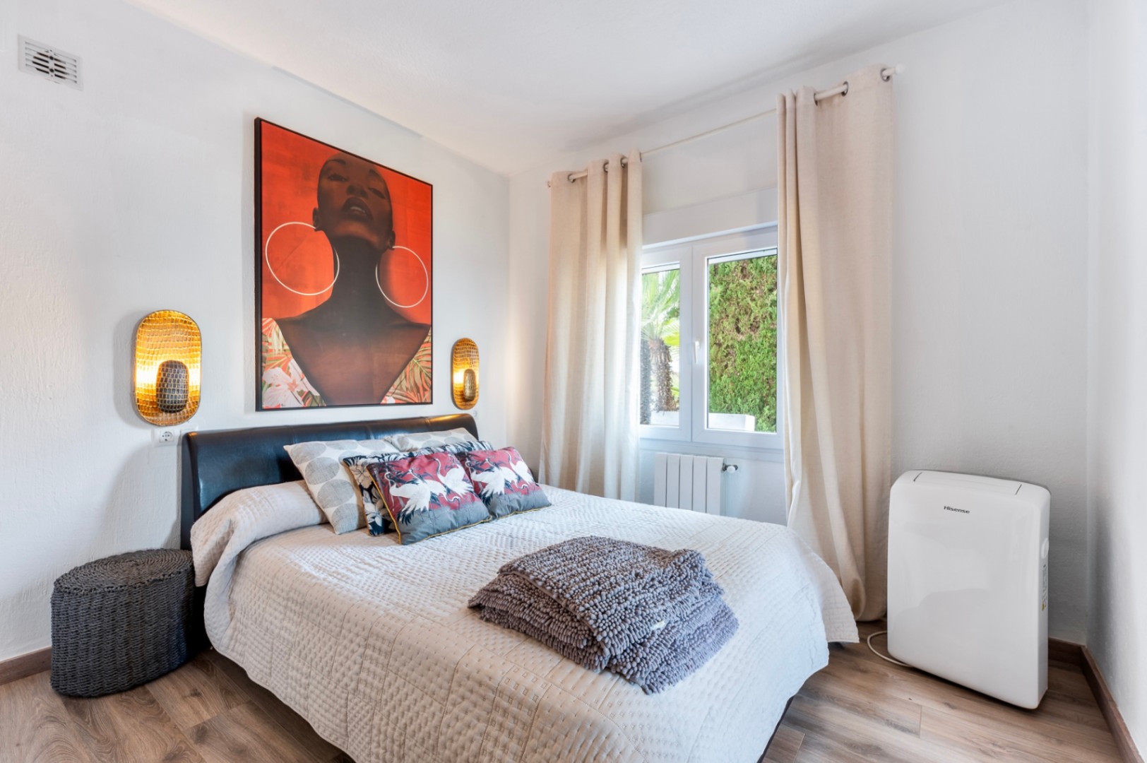 Chambre lumineuse avec parquet, tête de lit en cuir sombre et tableau d'art coloré. Comprend des appliques dorées, un radiateur et un climatiseur mobile.