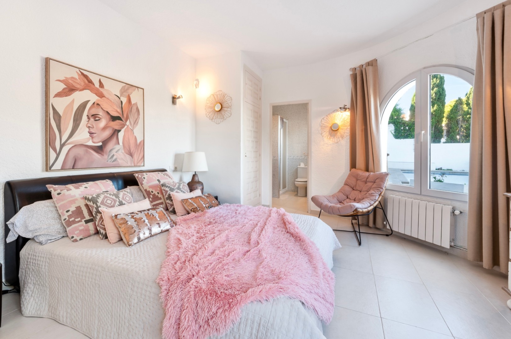 Chambre lumineuse avec salle de bain attenante, fenêtre en arc donnant sur l'extérieur et décoration moderne aux tons rosés avec sol en grès cérame.