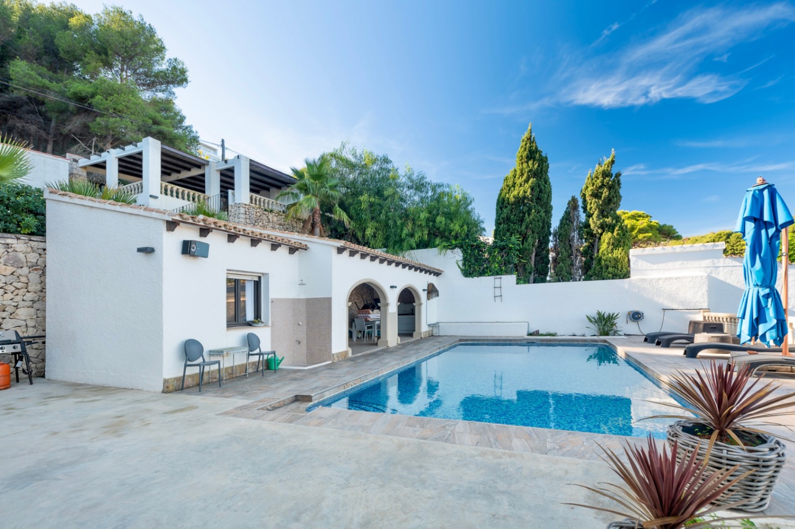 Espace piscine rectangulaire avec terrasse en grès, douche extérieure et porche à arches. L'environnement privé comprend des murs en pierre, des cyprès et une pergola supérieure.